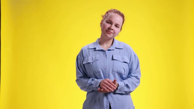 Young woman forcing polite smile while standing on yellow studio background. Concept of hidden annoyance, awkward social situation, uncomfortable reaction and polite communication.