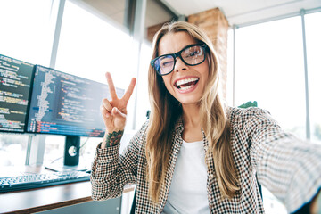 Young female programmer coder taking a happy selfie in a bright office workspace with dual monitors...