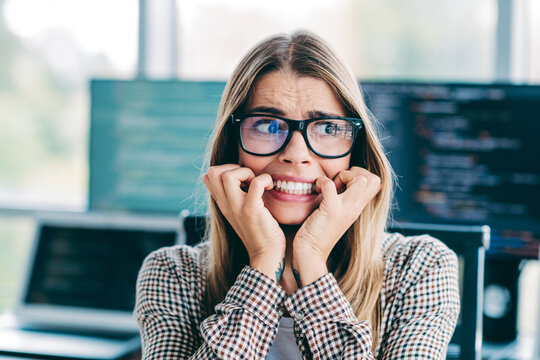 Stressed female programmer biting her nails while debugging complex code on multiple monitors in a modern open office, showing anxiety and deadline pressure