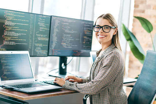 Female software developer smiling at desk, coding on laptop and dual monitors in a bright modern office workspace
