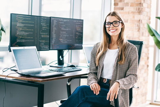Smiling female software developer at desk with dual monitors and laptop coding in a bright modern office