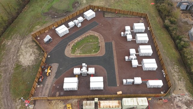 Aerial view of a fenced compound with multiple white container units and dark pathways, set against a backdrop of muted earth tones and sparse greenery, Burwell, United Kingdom.