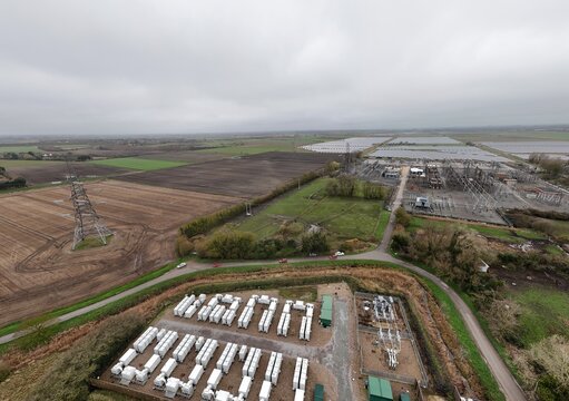 Aerial view of rows of white containers contrasting with green structures and a complex electrical substation under a cloudy sky, Burwell, United Kingdom.