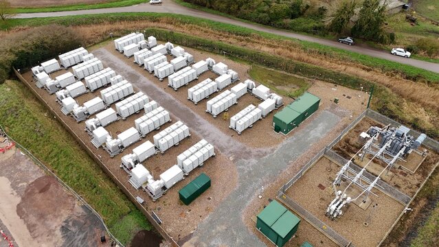 Aerial view of rows of gleaming white battery units nestled against the earth, contrasting with the green utility buildings, Burwell, United Kingdom.