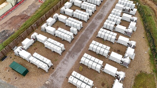 Aerial view of rows of white battery storage containers contrasting against the brown gravel and vibrant green foliage, Burwell, United Kingdom.