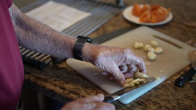 Senior man prepares garlic by mashing and mincing it in his home kitchen while following a recipe on a sunny afternoon.