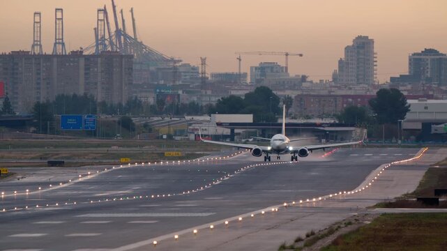 A passenger airplane with landing lights on makes a smooth touchdown on a wet runway. An industrial city and port are visible in the background during a hazy sunrise or sunset
