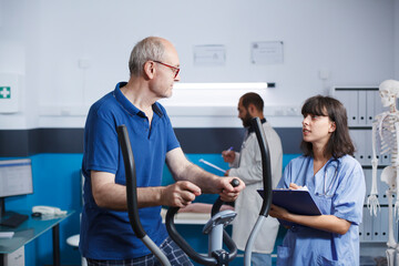 Fototapeta premium Elderly patient is seen receiving physiotherapy on stationary bike, assisted by female nurse dressed in blue scrubs. Senior man is getting rehabilitative care with help from health professional.