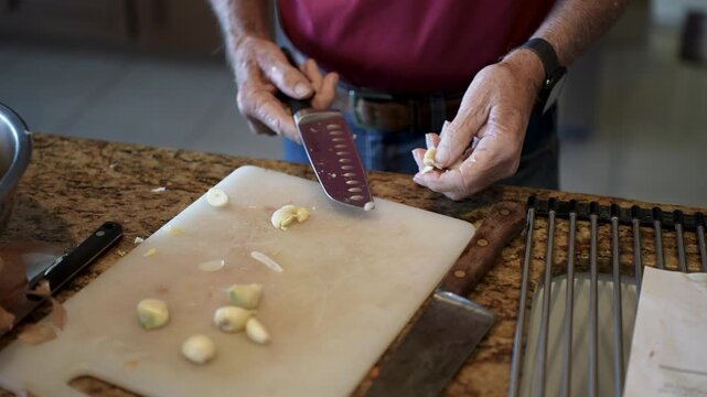 Elderly man is preparing garlic by mashing and mincing it while cooking in his home kitchen.