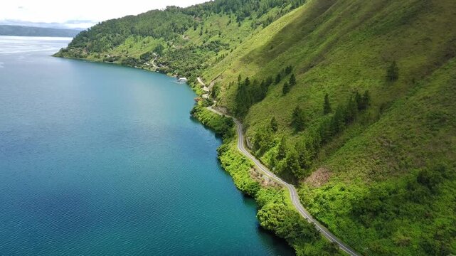 4K aerial drone footage flying parallel to hillside near Lake Toba, showing winding road, green hills, pine trees, and calm blue water with smooth cinematic motion