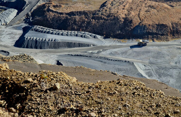 open pit mine quarry where raw materials are extracted. Mining equipment large yellow mining dump truck moving along paved road transporting excavated rock. Heaps of material stacked on top of piles o © Michal