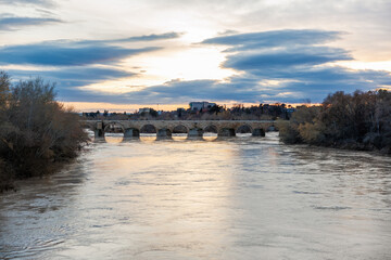 Obraz premium An old stone bridge with semicircular arches spans the Guadalquivir in Cordoba, Spain, as winter trees line the banks and golden reflections ripple near dusk under layered clouds.