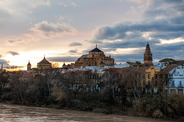 Riverside view in Cordoba, Spain, with Mezquita Catedral dome and bell tower above whitewashed houses, terracotta roofs, and winter bare trees at late afternoon. © True Pixel Art