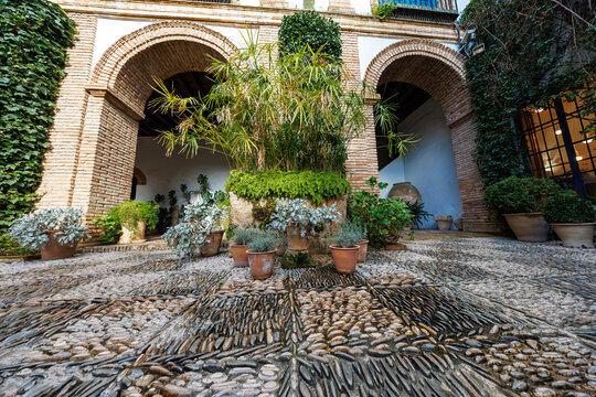 Traditional Andalusian patio in Cordoba shows pebble mosaic panels, Moorish arches, whitewashed walls, circular planter with ferns and papyrus, and a clay amphora.
