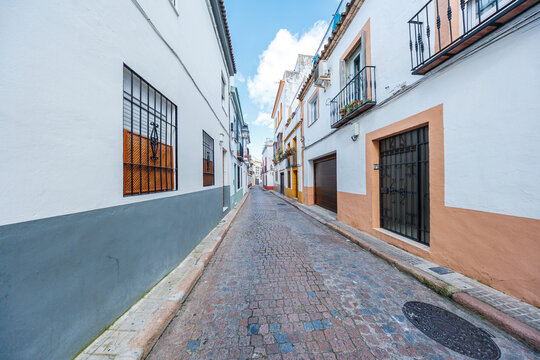 A cobblestone lane in Cordoba, Spain runs between whitewashed houses with wrought iron rejas and small balconies, tapering toward a church tower in midday light.