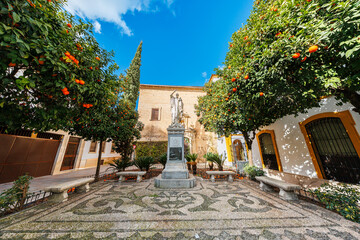 A stone statue on a plinth stands in a small Cordoba courtyard with benches, pebble mosaic paving, white facades with ochre trim, orange trees, and a tall cypress. © True Pixel Art