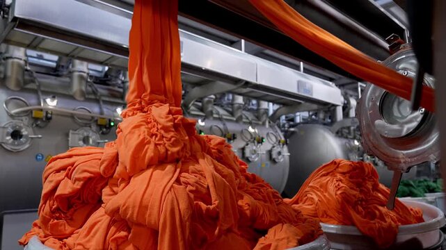 Bright orange fabric flowing from large dyeing machines in a textile factory. Professional industrial footage reflecting technical process, production control and large scale manufacturing.