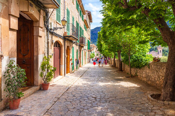 Fototapeta premium Tourists walking through a picturesque shaded street in Valldemossa village, Mallorca, with stone buildings and green shutters in the UNESCO Tramuntana region.