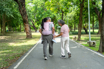 Obraz premium Back of Asian girl piggyback ride to senior man and walk together with senior woman along the road in public park and they look happy to relax with nature together.
