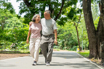 Fototapeta premium Asian senior man put his arm around senior woman and walk along the road in public park and they look each other with happiness during exercise and relax together.