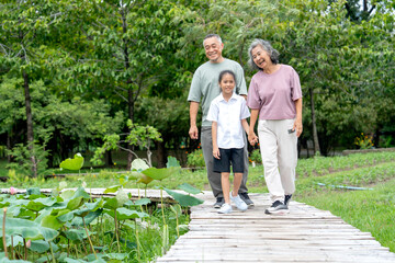 Obraz premium Asian senior man and woman walk on a wooden bridge that stretches across a field of lotus flowers together with the girl and they look happiness in public park with day light.
