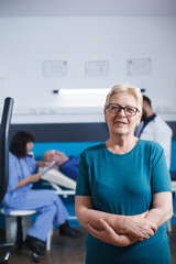Fototapeta premium Old woman in portrait waits to work out for physical treatment with arms crossed. Senior patient with muscular injury gets ready for leg physiotherapy on stationary bike while she looks at camera.