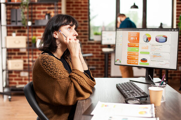Female employee sits at office desk having brainstorming session as desktop monitor displays market...