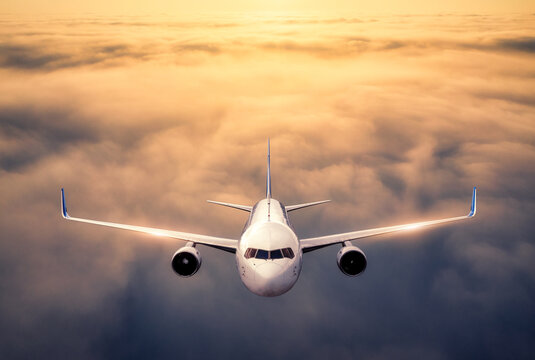 Front view of white commercial passenger airplane flying above clouds at sunset. Landscape with passenger aircraft, low clouds, orange sky. Aerial view. Aviation and air travel concept. Transportation