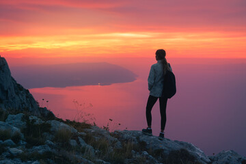 Fototapeta premium Silhouette of woman with backpack standing on rocky mountain peak at sunset in Croatia, overlooking the sea with colorful pink sky. Outdoor travel adventure. Girl hiker on mountain at twilight