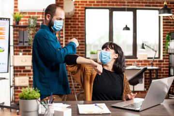 Fototapeta premium Businesswoman working at desk as male coworker greets her with elbow in brick wall office. Businessman and female colleague wearing face mask and maintaining safety in startup company.