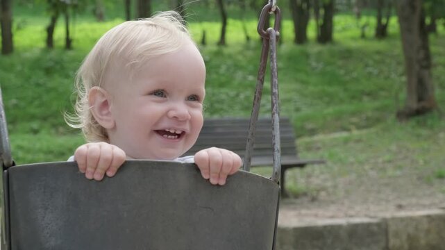  Closeup video of a small caucasian boy smiling while swinging on a swing on the kid's playground. Beautiful kid swinging on a swing and smiling on a nice summer day in the park. Slow motion.
