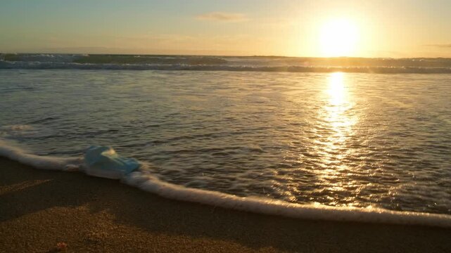 Low angle view of Ocean shore with a medical mask washing on shore by the waves with an orange sun on the horizon at sunset. Sandy beach with rolling waves with a masl during sunset. slow-motion.