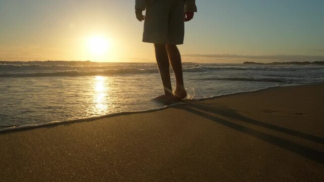 Happy young man walking into a sandy ocean beach on a sunny summer day. Young Male guy standing in the waves along the sandy coast on a sunny evening. A guy going to touch the seawater. slow motion.