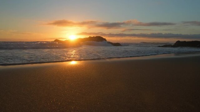 Sandy Ocean shore with rolling waves with an orange sun on the horizon at sunset. Sand beach with rolling waves hitting a big rock. Contemplation of the sea waves during sunset. Slow motion.
