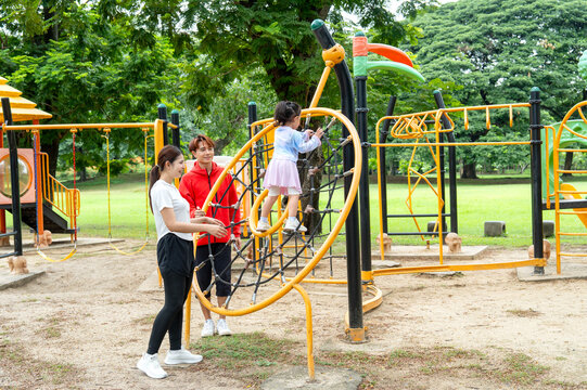Asian mother stand behide the little girl to take care during her child enjoy to play in the playground of public park with daylight.