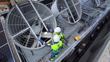 Two technicians in safety gear inspect large industrial cooling tower fans on a rooftop © FotoArtist