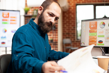 Fototapeta premium Closeup of bearded man seated at desk, focused on reviewing market analysis documents. Male manager looking at financial paperwork as computer displays graphs and analytics tools in brick wall office.