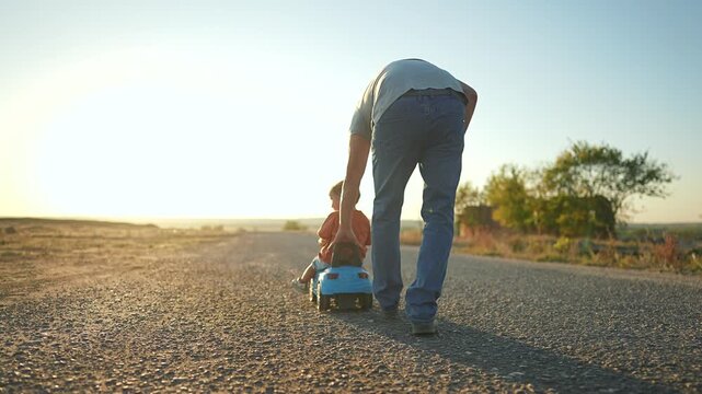 Father playing with child outdoors. Happy Child sits on toy car while father pushes. Playtime bonding between child and father. Summer day fun with car. Child enjoys ride in toy car with father.