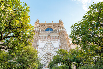 Fototapeta premium The Facade of Seville Cathedral rises behind orange trees in Seville, Spain. Visitors look upward at Gothic tracery, statues, and a large blue rose window in warm daylight.