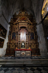 Obraz premium Baroque altarpiece dominates a side chapel at Seville Cathedral, Spain. Gilded wood frames saints and a veiled reliquary bust under Gothic vaults and spot lighting.