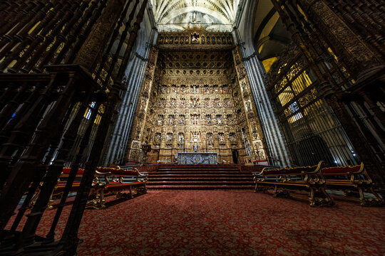 Seville, Spain interior shows the Capilla Mayor, an immense gilded retablo behind an altar, deep red carpeting, ornate choir stalls, and iron screens under low lighting.