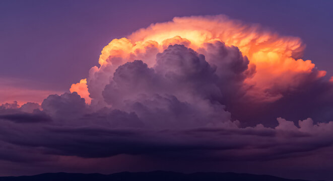 Towering cumulonimbus storm cloud illuminated by vibrant sunset light showcasing dramatic orange and purple atmospheric colors above a dark distant horizon line