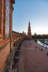 Fototapeta premium Late day view of Plaza de Espana in Seville shows semi circular arcade, patterned tile benches, south tower, canal, and visitors in soft early evening light.