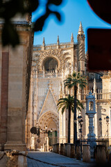 Seville, Spain, Seville Cathedral Gothic facade shows rose window, pointed arch, pinnacles, palm trees, lampposts, bell monument, chained posts in late afternoon light. © True Pixel Art