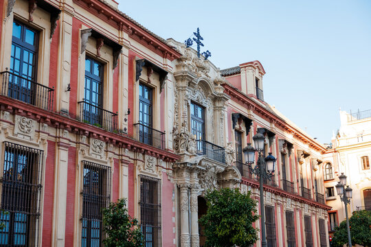 Archbishop's Palace Churrigueresque Portal, Seville, Spain. Street level view shows pink and cream pilasters, wrought iron balconies, carved entrance, cross, lampposts, and orange trees.