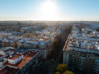 Obraz premium Late afternoon aerial over Seville residential grid and avenue. White facades and terracotta roofs line a straight route, long shadows, terraces, dishes, and tanks.