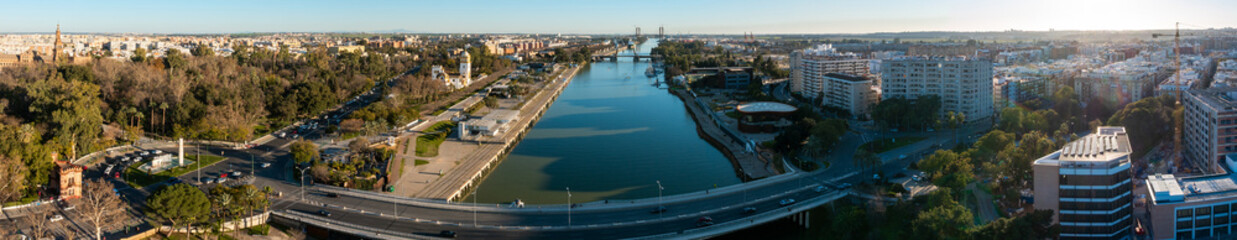 Naklejka premium Wide panorama shows Guadalquivir River in Seville, with Torre del Oro, Puente del Alamillo, curving bridge, multi lane roadway, moored boats, and Maria Luisa Park in late light.