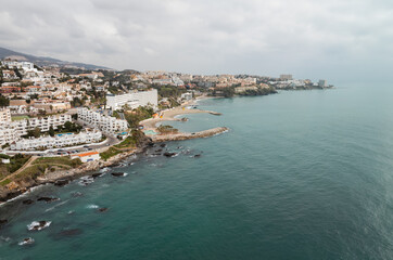 Aerial view shows Benalmadena coastline in Malaga with white villas, resort hotels, crescent beaches, stone breakwaters, turquoise water, and surf on dark rocks in soft late light.