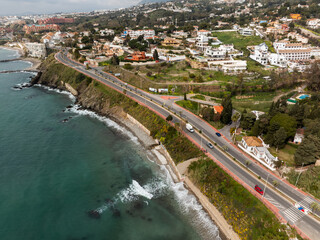 Aerial view of a curving cliffside highway with red sidewalks and palm lined barriers above turquoise water near Malaga, Costa del Sol, Spain, under midday light.