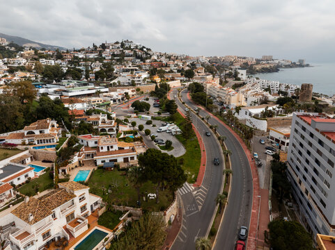 Aerial scene shows whitewashed villas, tiled roofs, courtyard pools, and palm lined avenue curving to the Mediterranean and rocky promontory in Spain under overcast light.